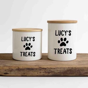 May include: Two white cylindrical ceramic canisters with light brown wooden lids. Each canister features the black text "LUCY'S" above a black paw print graphic and the word "TREATS" below it. The canisters are of different heights.