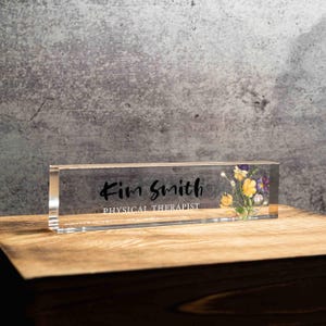 May include: A clear acrylic desk nameplate with the name "Kim Smith" and the title "PHYSICAL THERAPIST" in black script. The nameplate features a floral design with yellow and purple flowers. The nameplate is displayed on a wooden surface.