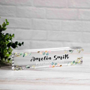 May include: Clear acrylic desk nameplate with the name "Amelia Smith" and the title "Psychologist" in black script. The nameplate is decorated with watercolor-style leaves in shades of green, pink, and peach.