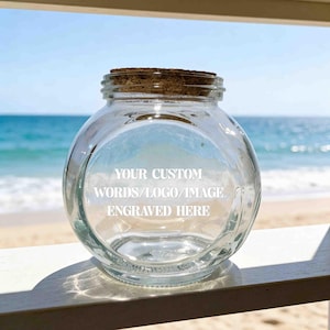 Könnte beinhalten: Ein klares, rundes Glasgefäß mit einem Korkstopfen. Auf der Vorderseite befindet sich eine weiße Gravur mit dem Text: YOUR CUSTOM WORDS/LOGO/IMAGE ENGRAVED HERE. Das Gefäß hat einen bauchigen Körper und einen kurzen, schmalen Hals.