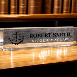 May include: A clear acrylic desk nameplate with the name "ROBERT SMITH" and the title "ATTORNEY AT LAW" in black lettering. A scale of justice and laurel wreath are on the left. The nameplate sits on a wooden desk, with law books in the background.