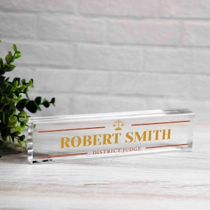 May include: Clear acrylic desk nameplate with gold lettering. The nameplate reads "ROBERT SMITH DISTRICT JUDGE" with a scale of justice symbol. The nameplate is rectangular and sits on a light wood surface.