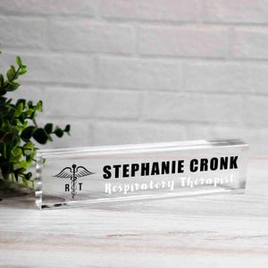 May include: A clear acrylic desk nameplate with the name "STEPHANIE CRONK" in bold black letters and "Respiratory Therapist" in a smaller, lighter font. A medical symbol is on the left. The nameplate is on a white wooden surface.
