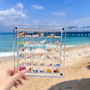 May include: A square clear acrylic display case with a grid of compartments containing various small seashells and colorful sea glass. The text "Dockweiler Beach, Playa Del Rey, CA 2026" is printed in white across the center of the transparent frame.