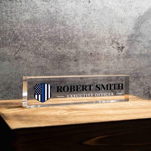May include: A clear acrylic desk nameplate with the name "ROBERT SMITH" and the title "EXECUTIVE OFFICER" in black text. A blue and white shield graphic is on the left side. The nameplate sits on a wooden surface.