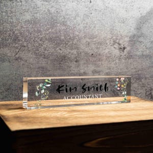 May include: A clear acrylic desk nameplate with the name "Kim Smith" and the title "Accountant" in black script. The nameplate is decorated with floral accents in green, pink, and yellow. The nameplate is on a wooden surface.