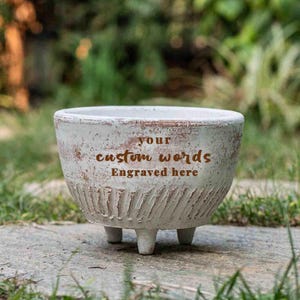 Op de afbeelding: Een ronde, gebroken witte keramische plantenbak met een verweerde afwerking. De buitenkant heeft een textuur met ribbels aan de onderkant en de bruine tekst "your custom words Engraved here". De plantenbak rust op drie korte, stevige pootjes.