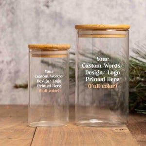 May include: Two clear cylindrical glass storage jars with light brown wooden lids. Each jar features white and orange text that reads: Your Custom Words/ Design/ Logo Printed Here (Full color). The jars are empty and have a smooth glass finish.