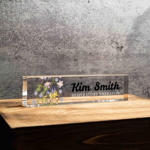 May include: A clear acrylic desk nameplate with the name "Kim Smith" and the title "Respiratory Therapist" in black lettering. The nameplate features a floral design with purple, yellow, and blue flowers.