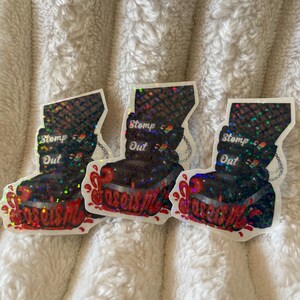 May include: Three black and iridescent boot-shaped stickers with the words "Stomp Out Fascism" and "Stomp Out" printed on them. The boots are dripping with red paint.
