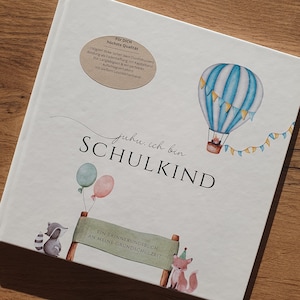 Könnte beinhalten: Ein weißes Buch mit dem Text "juhu. ich bin SCHULKIND" und einer Illustration eines Heißluftballons. Das Buch trägt den Titel "Ein Erinnerungsbuch an meine Grundschulzeit".
