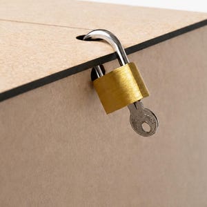 Puede incluir: Primer plano de una caja de madera beige con borde negro, asegurada con un candado y una llave dorados. La llave est&aacute; insertada en la cerradura, lo que sugiere que la caja est&aacute; cerrada. La caja tiene una ranura en la parte superior.