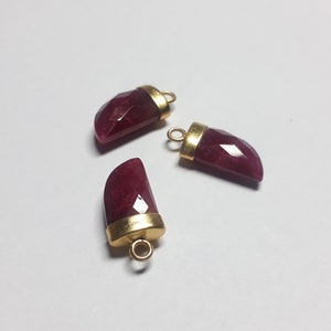May include: Three faceted, horn-shaped pendants with a deep red hue. Each pendant has a gold-colored metal cap with a small loop for attaching to a chain or other jewelry component. The pendants are arranged on a white surface.