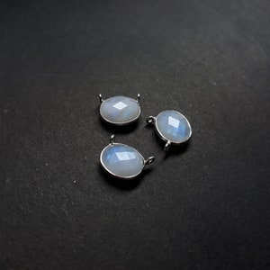 May include: Three oval-shaped moonstone pendants with silver-colored settings. Each pendant has a faceted surface that reflects light, creating a shimmering effect. The pendants are displayed on a dark background.