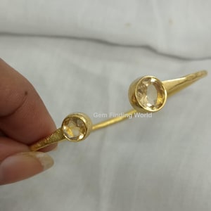 Könnte beinhalten: Ein goldfarbenes Armband mit zwei runden, hellgelben Edelsteinen. Das Armband hat eine strukturierte Oberfläche und ein schlichtes, elegantes Design. Die Edelsteine sind in einer Zargenfassung gefasst.