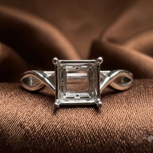 Puede incluir: Un engaste de anillo de metal plateado con una cabeza cuadrada de garras diseñada para sostener una piedra preciosa. La banda presenta un diseño trenzado y calado en ambos lados, que conduce al engaste central de cuatro garras con un centro hueco.