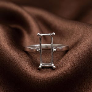 May include: A silver-toned metal ring setting featuring a vertical rectangular head with four prongs at the corners, designed to hold a gemstone. The band is smooth and polished, showcasing a clean, structural design for jewelry assembly.