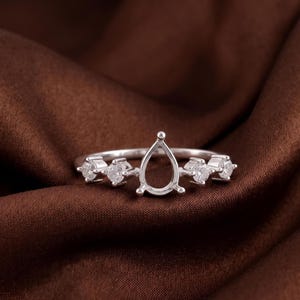 May include: A silver-toned ring featuring a central pear-shaped prong setting designed to hold a gemstone. The band is accented with two small round clear stones on each side of the central setting, held in place by four-prong metal mounts.