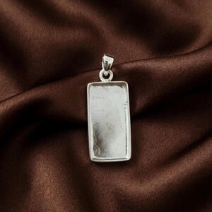 May include: A rectangular silver pendant tray with a smooth, flat interior surface and a raised border. The pendant features a small, circular silver bail at the top, designed for attachment to a necklace chain or cord, showcasing a metallic finish.