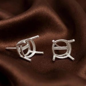 May include: Two silver-colored earring settings with a post and a four-prong design. The settings are empty, ready for gemstones. The earrings are displayed on a brown satin fabric.