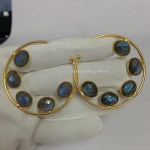 Può includere: Un paio di orecchini a cerchio color oro, ciascuno ornato da sette pietre preziose ovali blu-grigio iridescente. Gli orecchini sono esposti su una superficie bianca.