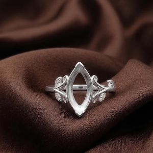 May include: A silver-toned ring setting featuring a central marquise-shaped bezel designed to hold a gemstone. The band is adorned with detailed leaf motifs extending from the sides of the central setting, creating a nature-inspired metal structure.