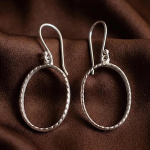 May include: A pair of silver oval hoop earrings. The earrings have a textured, rope-like design around the oval. The ear wires are simple hooks. The earrings are displayed on a brown fabric background.
