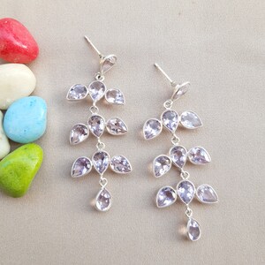 May include: A pair of silver-toned dangle earrings. Each earring features a cascading design of light purple, teardrop-shaped gemstones. The earrings are displayed on a neutral-colored surface, with colorful stones in the background.
