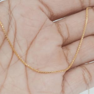 May include: A delicate gold-colored box chain necklace. The chain is composed of small, square links, creating a subtle, yet elegant design. The necklace is shown against a neutral background.