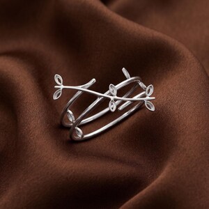 Puede incluir: Un anillo de tono plateado con un diseño cruzado que imita la apariencia de una rama delgada. La banda está adornada con pequeños motivos de hojas de marco abierto distribuidos a lo largo de los tallos metálicos, creando un aspecto delicado.