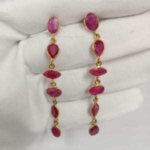 Peut inclure: Une paire de boucles d'oreilles pendantes dorées avec des pierres précieuses roses ovales et en forme de goutte. Les boucles d'oreilles ont un design de chaîne et de clou, avec plusieurs pierres précieuses en cascade. Sur fond blanc.