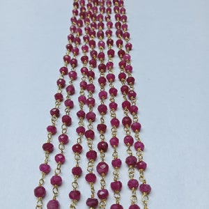 May include: Close-up of multiple strands of faceted, deep pink gemstones strung on delicate gold-colored chains. The beads are evenly spaced, creating a repeating pattern. The background is a plain white surface.