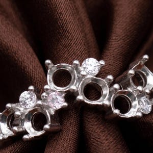 May include: A silver ring setting with multiple open bezels, each designed to hold a gemstone. The ring is adorned with small, clear, round-cut stones. The setting is displayed against a dark brown fabric background.