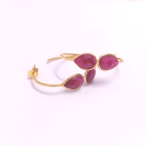May include: Gold-coloured hoop earrings with teardrop-shaped, faceted, red gemstones. The earrings have a post-and-clutch closure. The gemstones are arranged in a cluster design, adding a touch of elegance to the jewellery.