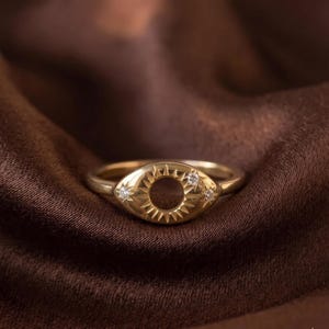 May include: A gold ring featuring an eye-shaped centerpiece with a central circular cutout surrounded by engraved sunburst rays and three small, clear, round-cut stones set into the metal surface on the left and right sides of the design.