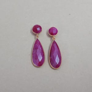 Op de afbeelding: Een paar goudkleurige oorbellen met een ronde, magenta steen aan de bovenkant en een druppelvormige, gefacetteerde, magenta steen eronder. Het eenvoudige en elegante ontwerp is geschikt voor verschillende gelegenheden.