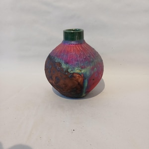 May include: A small, round ceramic vase with a narrow neck and a dark green rim. The body features a textured surface with a vibrant gradient of pink, purple, and blue, transitioning into a dark, mottled brown patch with a rough, organic appearance.