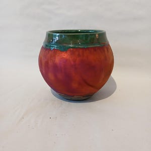 May include: A round ceramic pot with a wide, curved body and a short, narrow neck. The exterior features a vibrant, iridescent orange and red finish with a subtle vertical ribbed texture, while the rim is glazed in a deep, glossy forest green.
