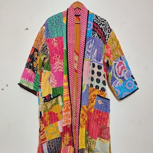 以下が含まれることがあります： 鮮やかな色のパッチワークの着物風ローブ。ピンク、イエロー、グリーン、ブルー、オレンジの色合いで、花柄、幾何学模様、抽象模様がミックスされています。長袖でゆったりとしたフィット感で、黒いトリムが付いています。