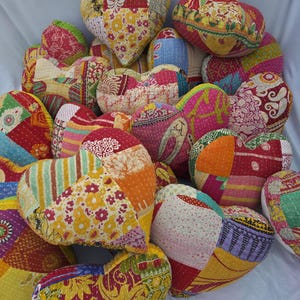 Può includere: Una collezione di cuscini a forma di cuore di varie dimensioni. I cuscini sono realizzati in tessuto patchwork in colori vivaci come rosso, giallo, arancione e rosa. Ogni cuscino ha un motivo unico, creando un mix visivamente accattivante ed eclettico. I cuscini sono disposti su una superficie bianca.