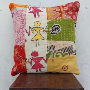 May include: Square patchwork pillow with a variety of fabrics in red, pink, green, and yellow. The pillow features hand-stitched details and a central panel with stick figure illustrations. The pillow is placed on a wooden stool.