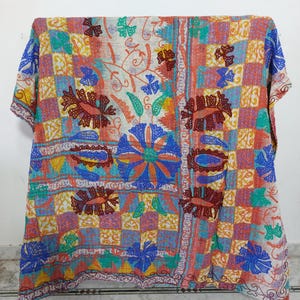 May include: A colorful, patterned textile with floral and geometric designs. The fabric features a patchwork of squares in shades of blue, orange, yellow, and green, with embroidered flowers and leaves. The textile is draped over a wooden frame.
