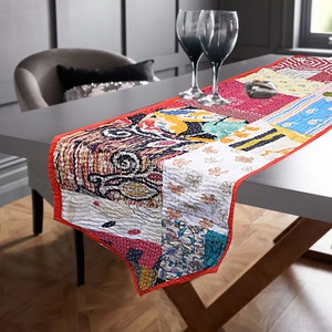 May include: A colorful patchwork table runner with a red border. The runner is made of different fabrics with various patterns, including floral, paisley, and geometric designs. It is laid on a white table with two wine glasses.