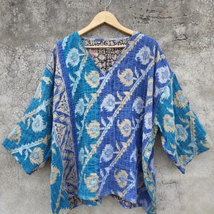 Könnte beinhalten: Ein blaues, türkisfarbenes und beigefarbenes Oberteil mit Muster, V-Ausschnitt und Dreiviertelärmeln. Das Oberteil ist aus einem gesteppten Stoff gefertigt und hängt an einem Holzbügel vor einer grauen Betonwand.