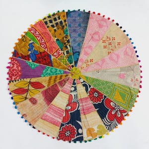 May include: Circular patchwork textile with a variety of patterned fabrics in pink, blue, yellow, and green. The edges are trimmed with colorful pom-poms. The design is a vibrant, handcrafted piece.