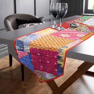 May include: A colorful patchwork table runner with a red border. The runner is made of different fabrics with various patterns and colors, including pink, blue, yellow, and green. The runner is laid out on a table with two wine glasses.