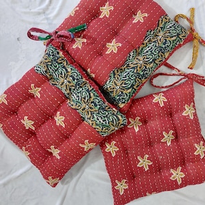 Puede incluir: Cojines de asiento rojos con estampados florales y costuras decorativas. Los cojines tienen forma cuadrada con un panel central estampado en contraste. Los cojines están atados con lazos de tela.