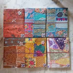 May include: A collection of six handmade, patchwork notebooks. Each notebook features a unique design with vibrant colors like orange, blue, yellow, and purple. The patchwork is adorned with floral and geometric patterns, and some have decorative embellishments.