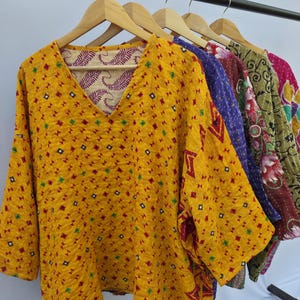 May include: A row of colorful, long-sleeved tops hanging on wooden hangers. The tops feature various patterns and colors, including yellow with geometric designs, purple, and floral prints. The tops have a V-neck design.
