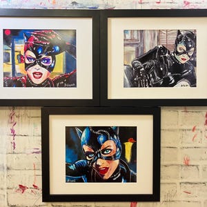 May include: Three framed watercolor paintings of Catwoman. Each painting features a different pose of the character, with a focus on her iconic mask and costume. The artwork is set against a white brick wall.
